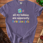 Purple t-shirt with text "all my hobbies are apparently forbidden spells" and cartoon book graphic.