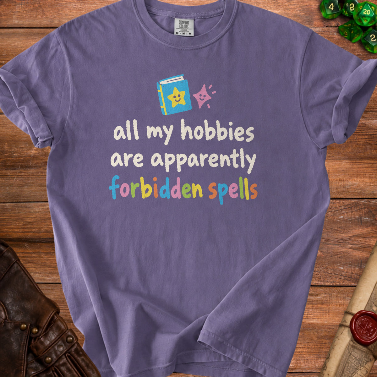 Purple t-shirt with text "all my hobbies are apparently forbidden spells" and cartoon book graphic.