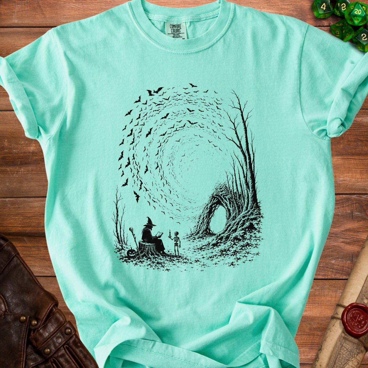 Mint green t-shirt with black graphic of witch, skeleton, and bats in a swirling vortex.