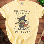 Yellow t-shirt with a goblin witch holding a cleaver and text "The Horrors Persist- But So Do T".