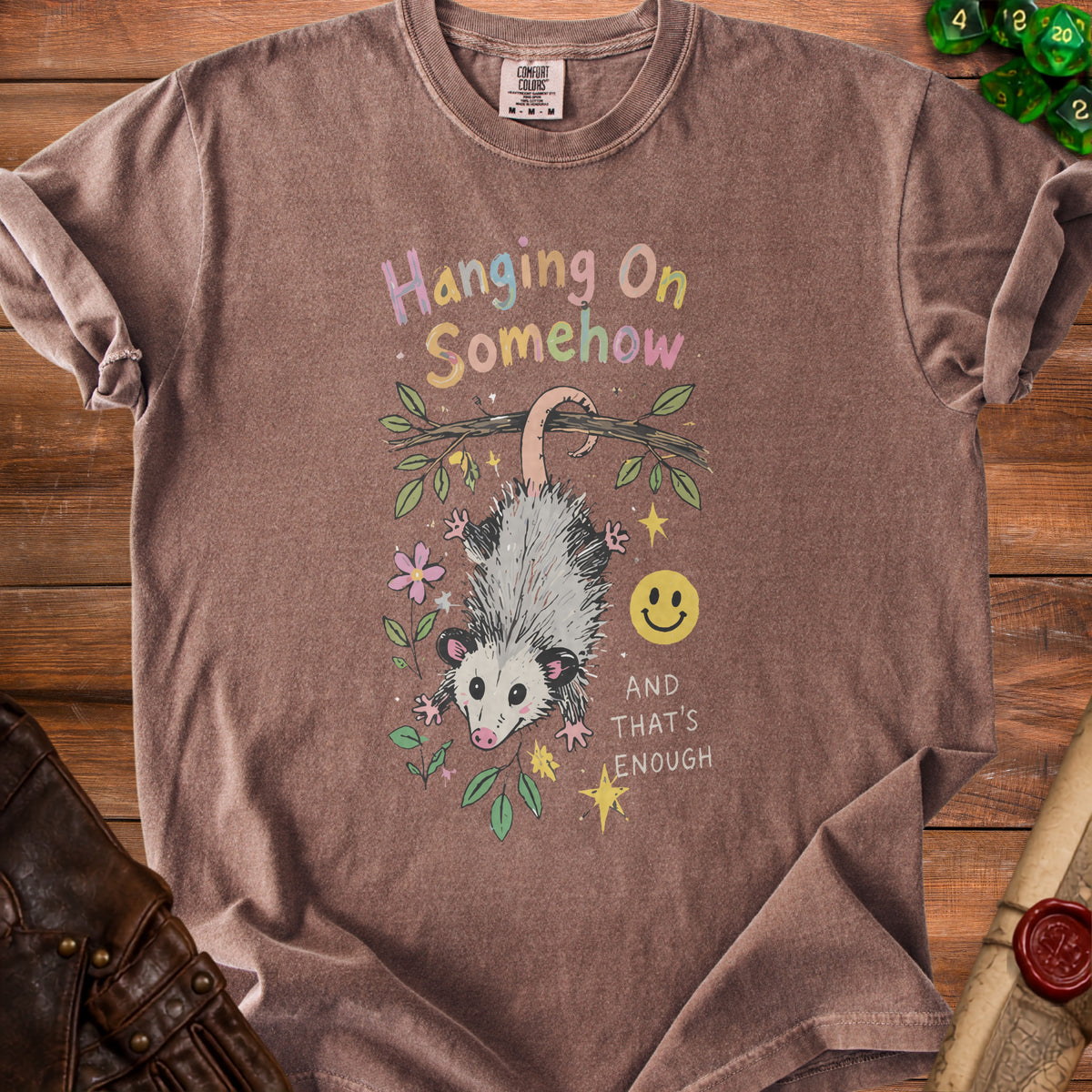 Brown t-shirt with opossum hanging from branch, text "Hanging On Somehow" and smiley face.