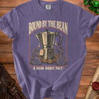 Purple t-shirt graphic with a moka pot, coffee beans, robed figures, and text "Bound By The Bean" and "A Dark Roast Pact".