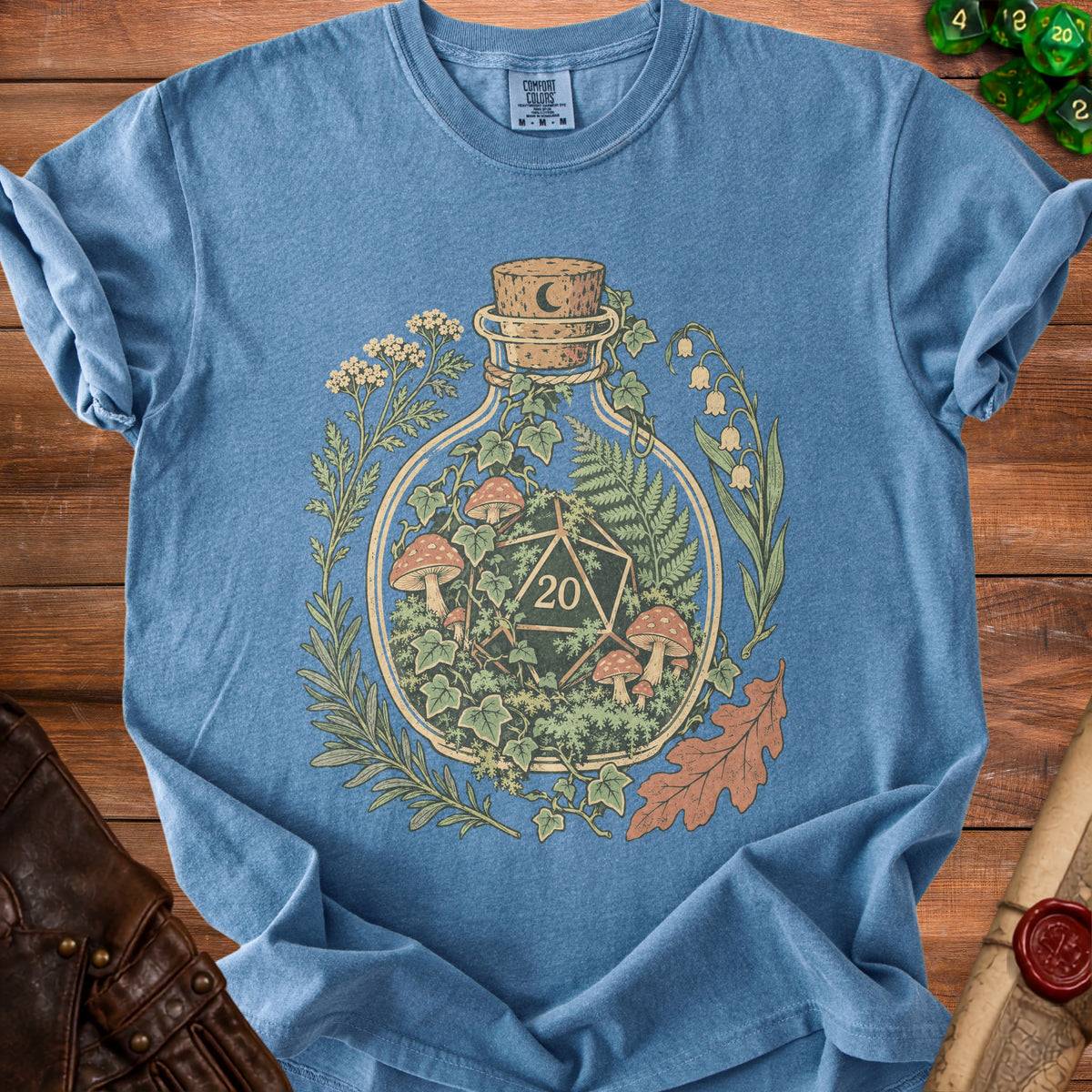 Blue t-shirt with a circular design of a bottle containing a D20 die, mushrooms, and plants.