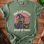Green t-shirt with a goblin wizard illustration and text "Bad at Spells Good at Vibes."