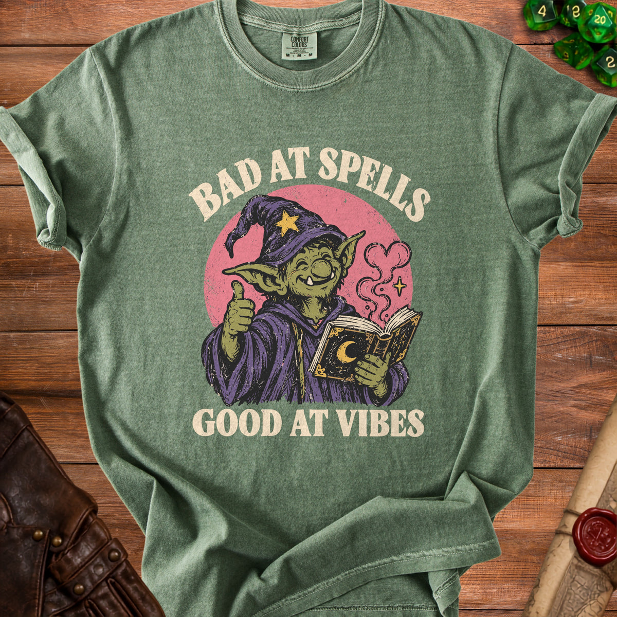 Green t-shirt with a goblin wizard illustration and text "Bad at Spells Good at Vibes."