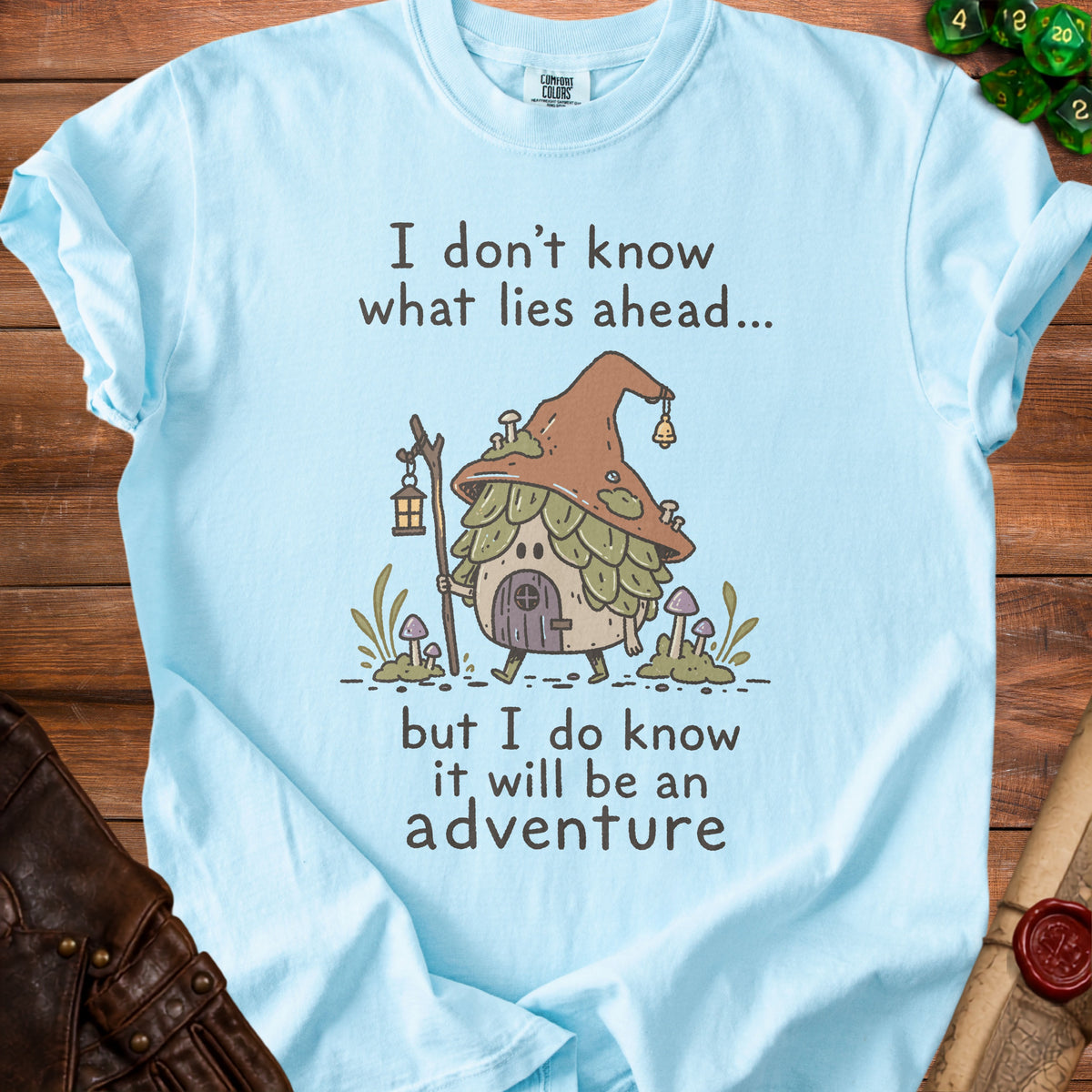 Light blue t-shirt with a drawing of a mushroom house, lantern, and the text "I don't know what lies ahead... but I do know it will be an adventure".