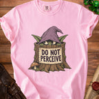 A pink t-shirt featuring a green goblin in a wizard hat hiding in a tree stump with text reading DO NOT PERCEIVE.