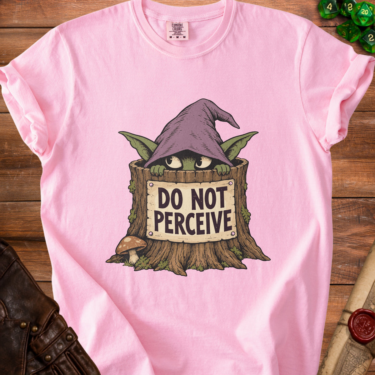 A pink t-shirt featuring a green goblin in a wizard hat hiding in a tree stump with text reading DO NOT PERCEIVE.