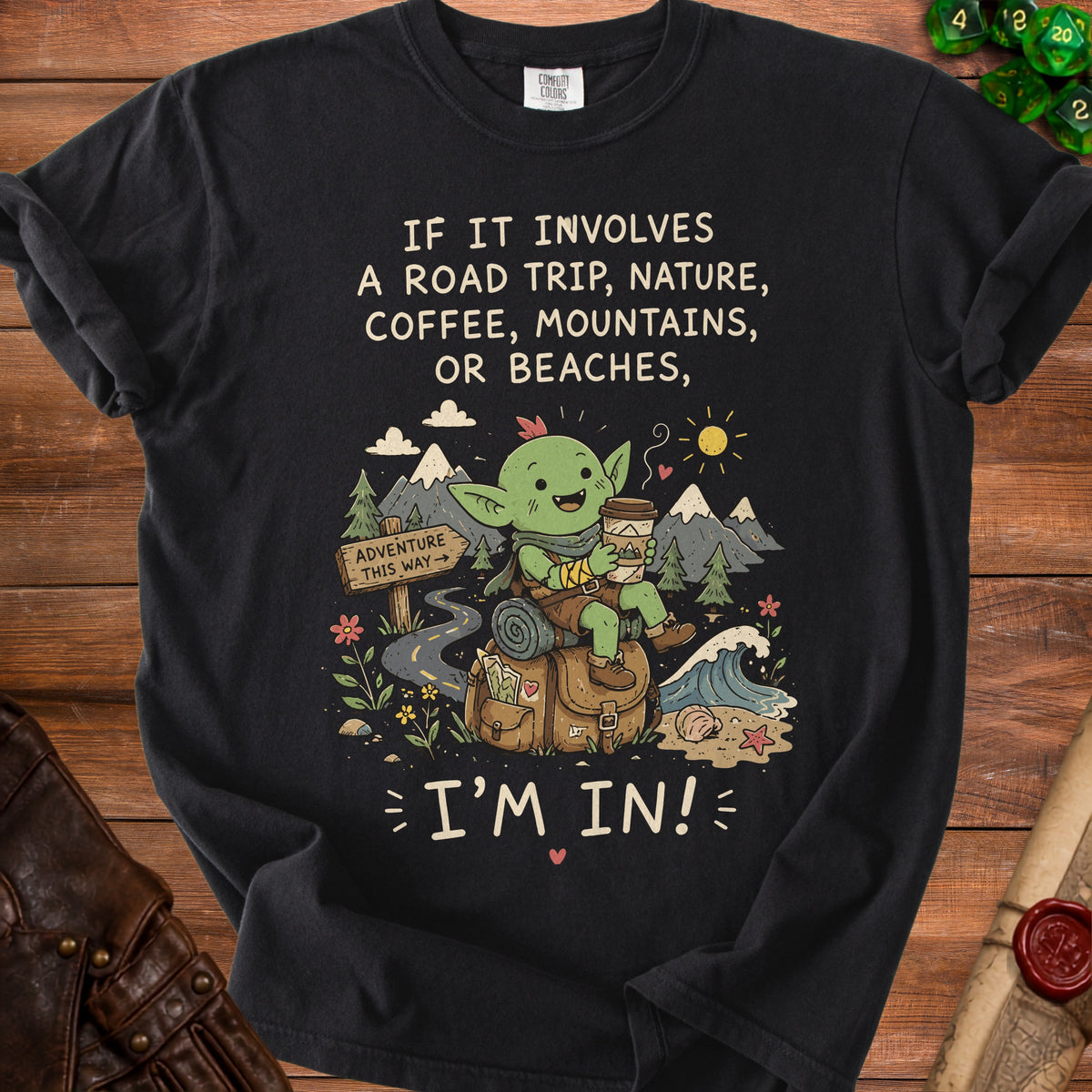 Black t-shirt with cartoon creature, mountains, and text "IF IT INVOLVES A ROAD TRIP, NATURE, COFFEE, MOUNTAINS, OR BEACHES, I'M IN!"