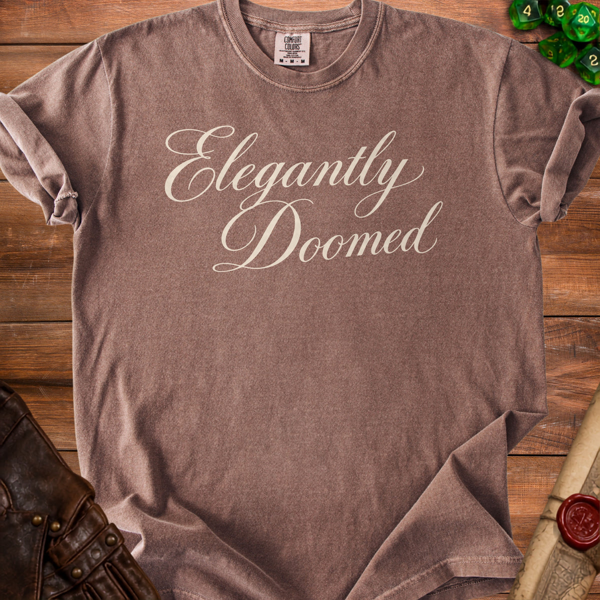 Brown t-shirt with "Elegantly Doomed" text, on a wooden surface.