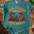 Teal t-shirt featuring a witch reading, a knight feeding a small dragon, and the text God Forbid Women Have Hobbies.