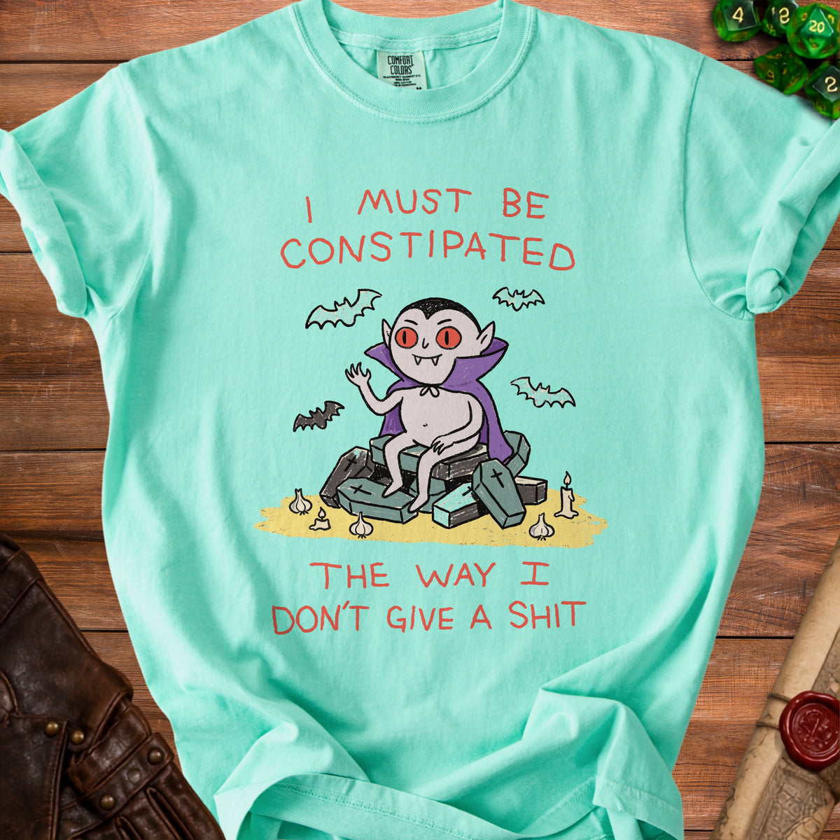 Mint t-shirt with cartoon vampire sitting on coffins, surrounded by bats, garlic, and a candle, text reads "I must be constipated the way I don't give a shit."