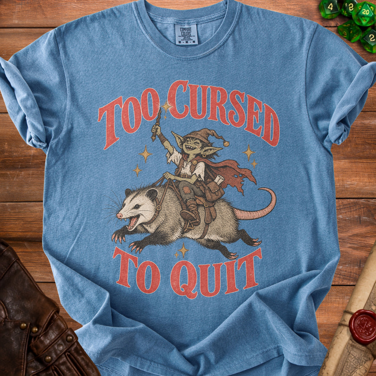 Blue t-shirt with goblin riding opossum and text "Too Cursed to Quit".