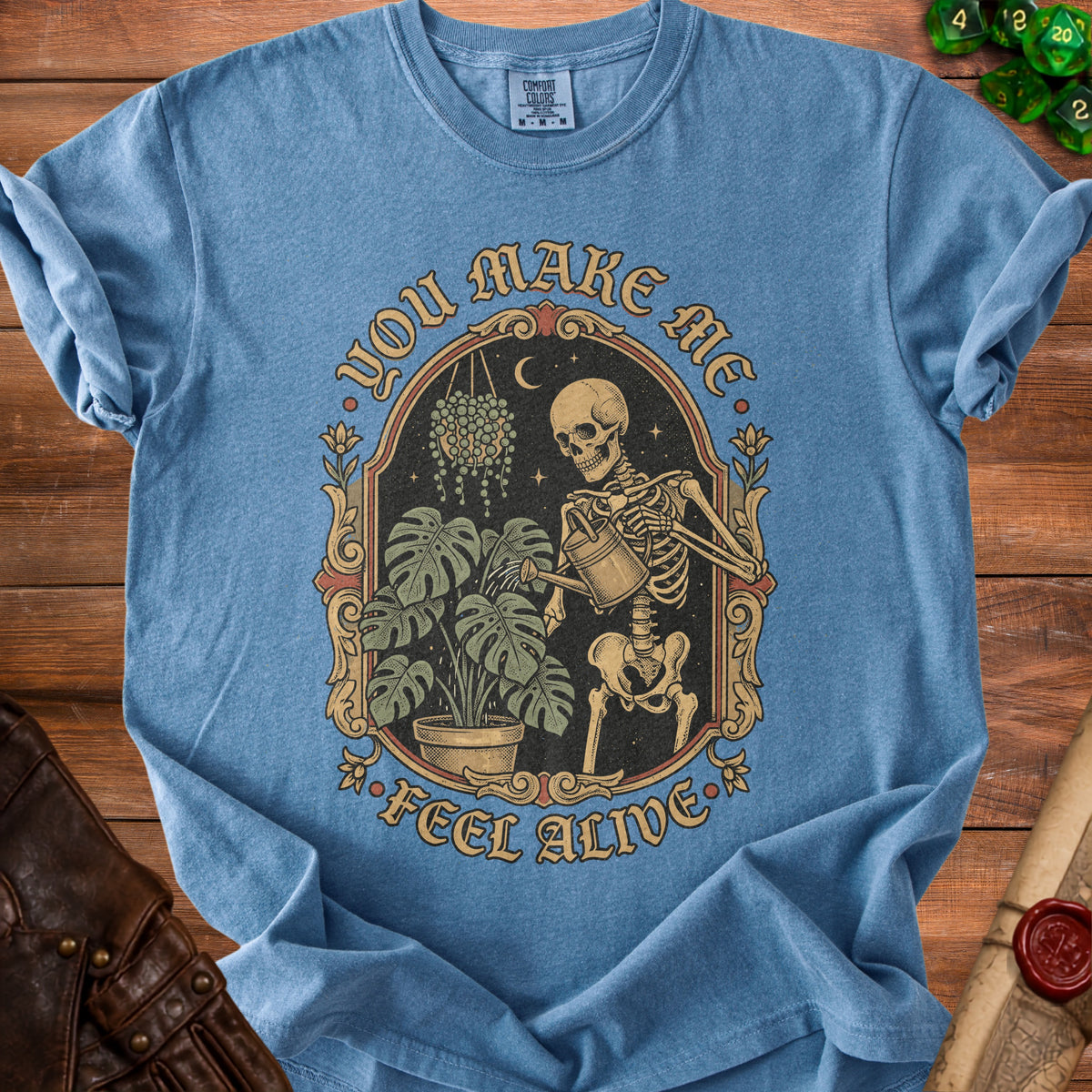 Blue t-shirt with skeleton watering plants graphic and "You Make Me Feel Alive" text.