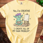 Yellow t-shirt with cartoon creature, easel, and text: "Yes, I'm CREATIVE I CREATE ALL OF MY OWN PROBLEMS".