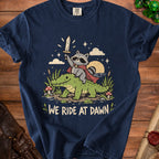 T-shirt graphic of a raccoon riding an alligator with the text "We Ride At Dawn."