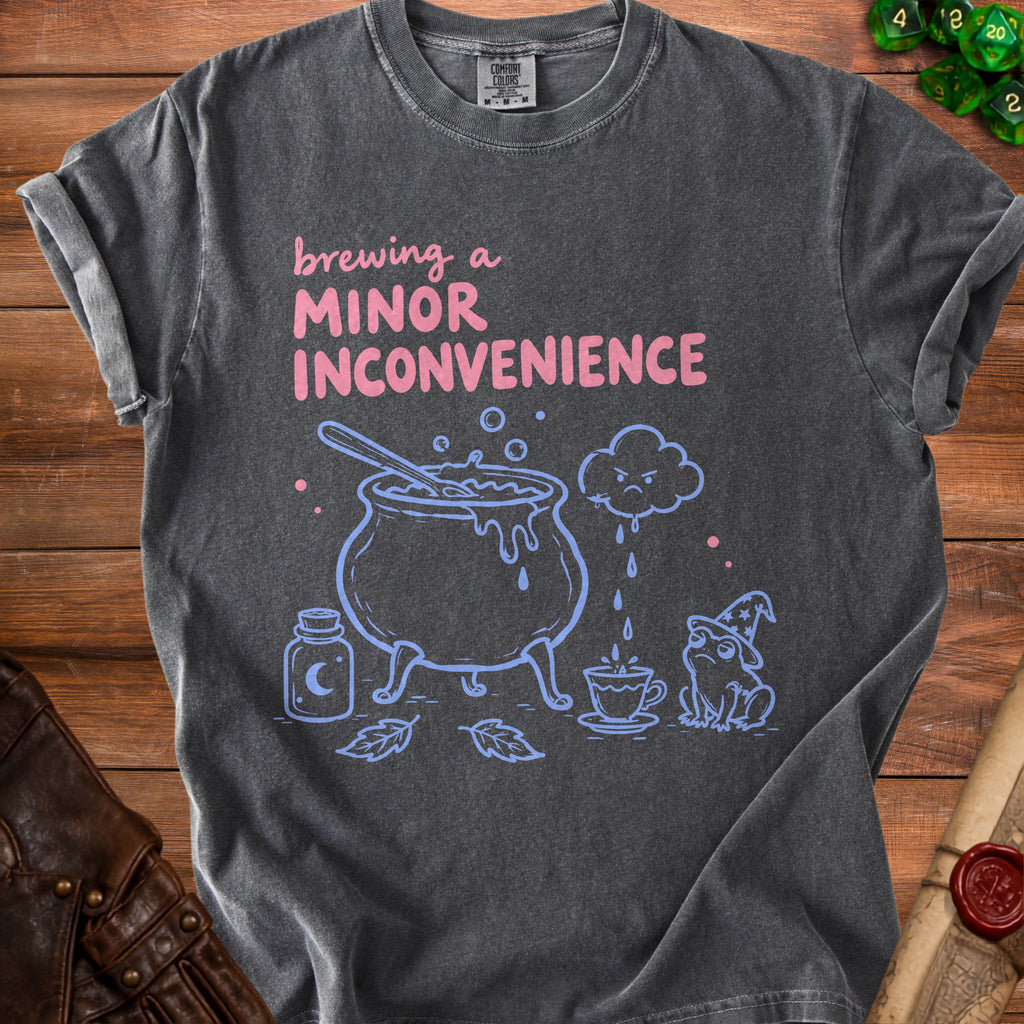 Gray t-shirt with pink text "brewing a minor inconvenience" and blue witch's cauldron, frog, and potion.