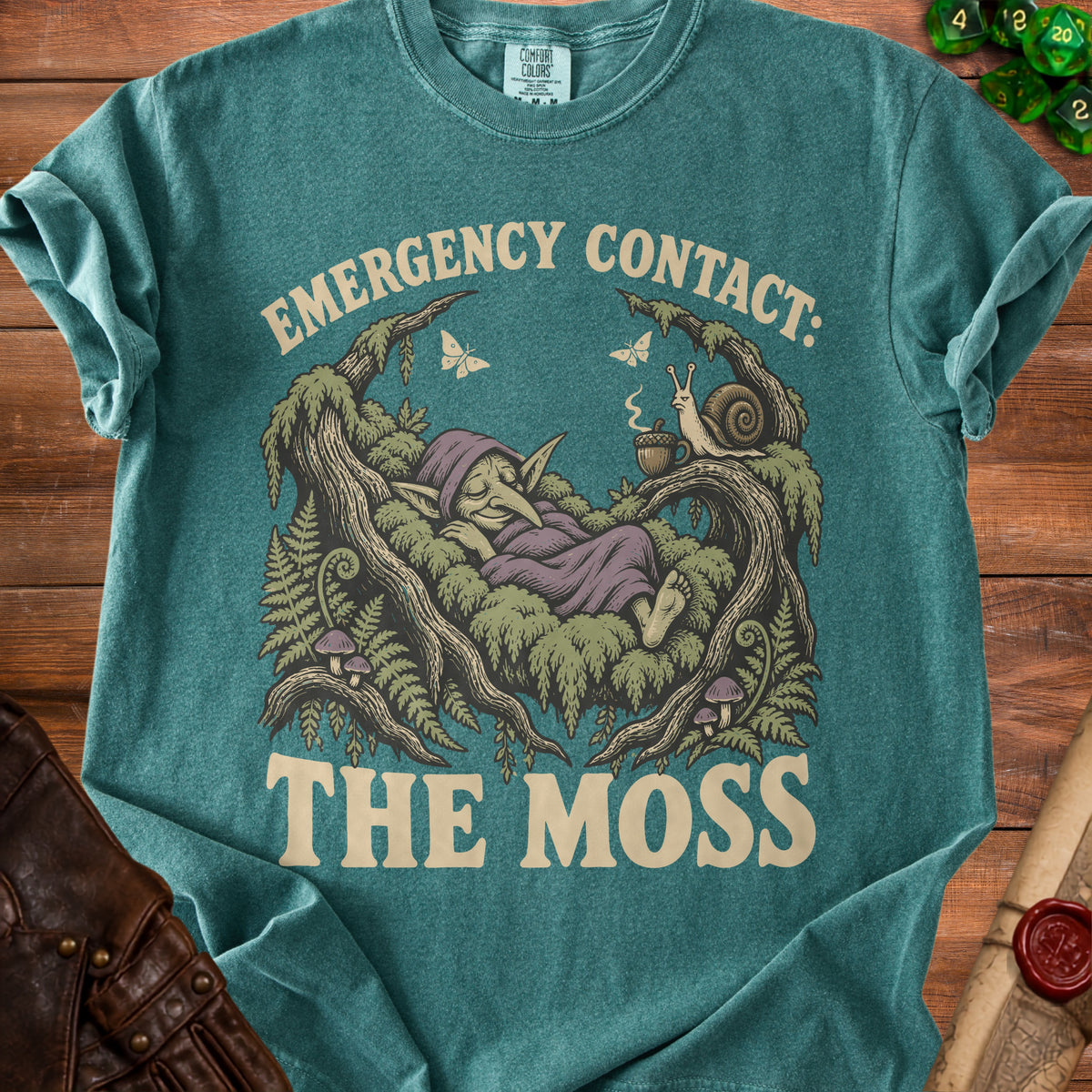 Teal t-shirt with a gnome sleeping amongst mossy trees, text reads "EMERGENCY CONTACT: THE MOSS".