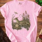 A pink t-shirt featuring a detailed illustration of a dragon resting near water with a twenty-sided die.