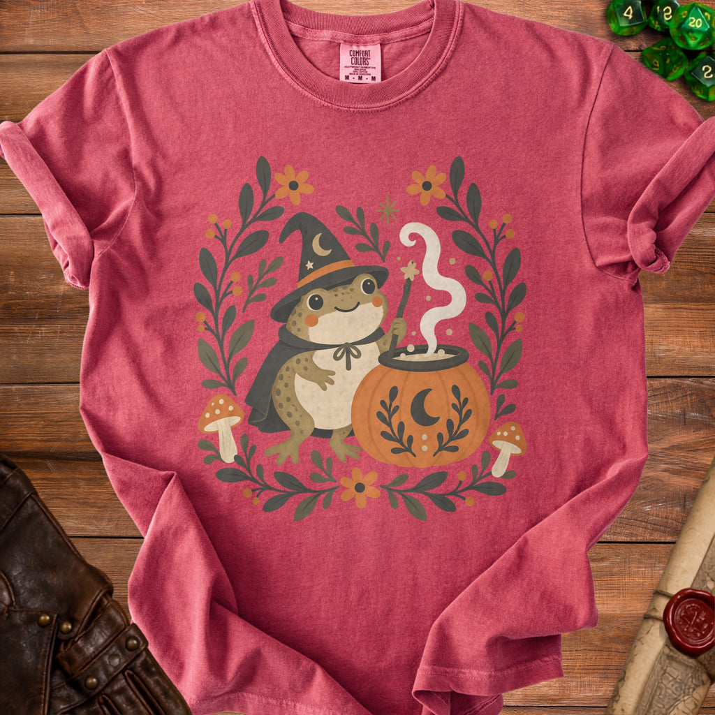 T-shirt with a cartoon frog wizard stirring a cauldron within a floral border.