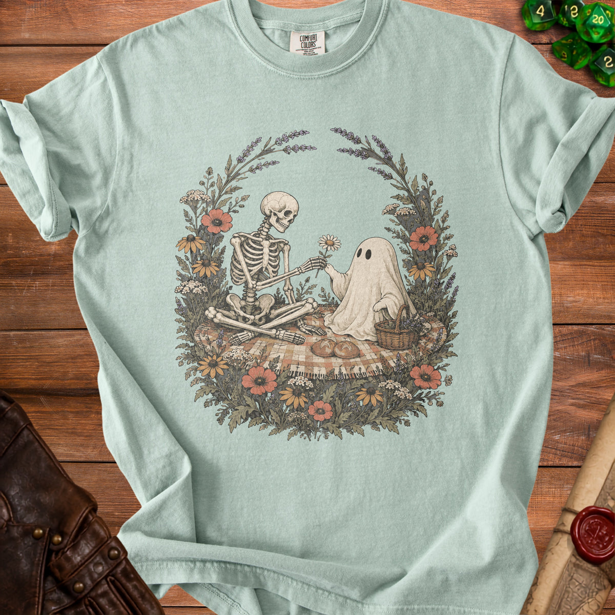 Sage green t-shirt featuring a vintage-style graphic of a skeleton and a ghost having a picnic surrounded by flowers.