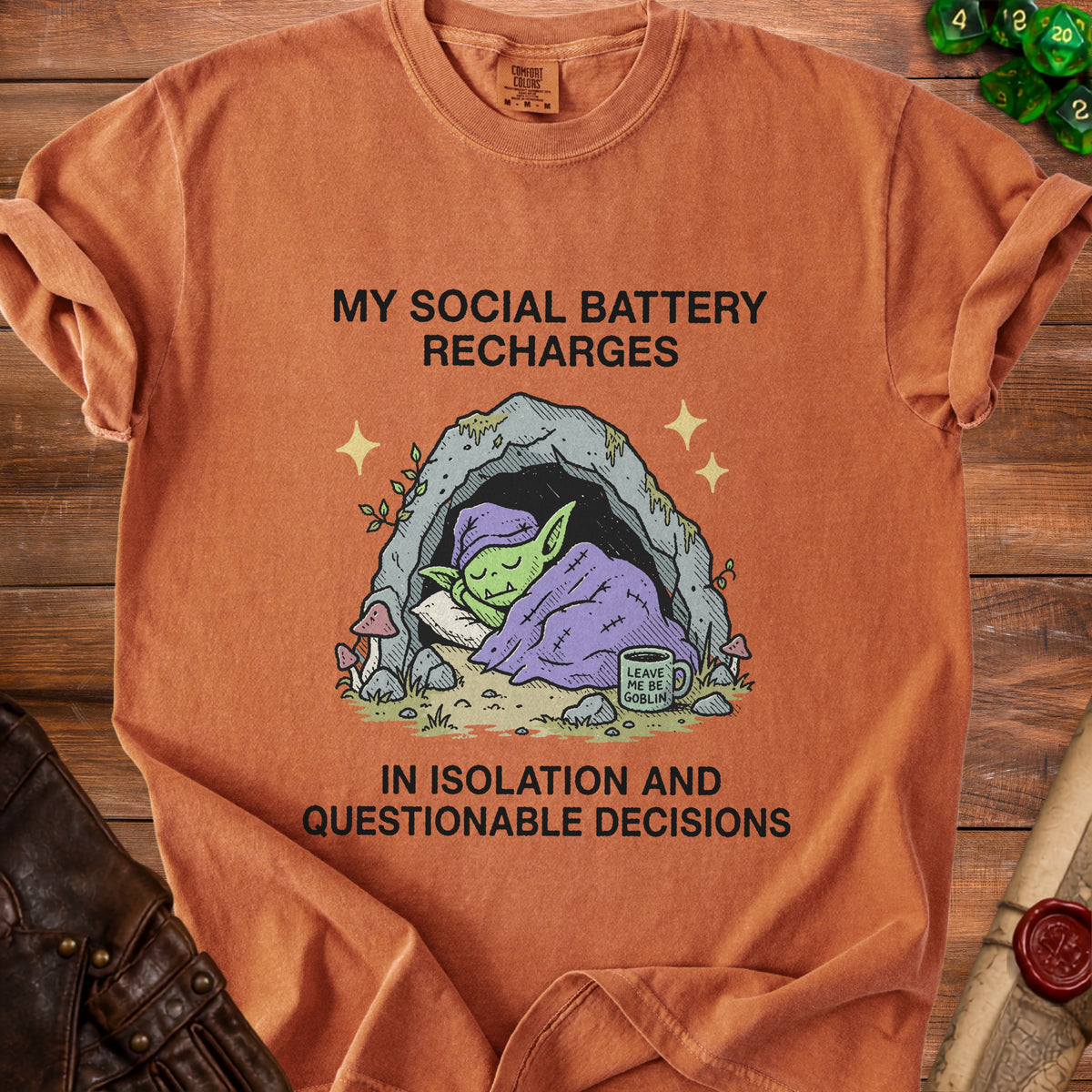 Orange t-shirt with a graphic of a goblin sleeping in a cave with text "My social battery recharges in isolation and questionable decisions."