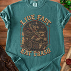 Green t-shirt with "Live Fast Eat Trash" text and cartoon creatures in a cart.