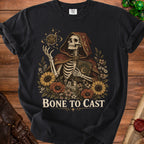Black t-shirt with a skeleton in a hooded robe holding a book and a d20 dice.