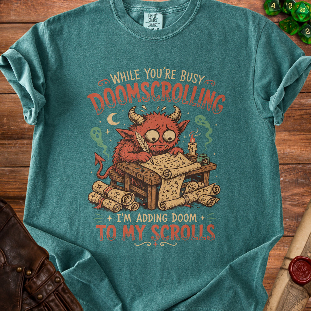 Green t-shirt with a red devil writing on scrolls, reading "While you're busy doomscrolling, I'm adding doom to my scrolls."