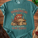 Green t-shirt with a red devil writing on scrolls, reading "While you're busy doomscrolling, I'm adding doom to my scrolls."
