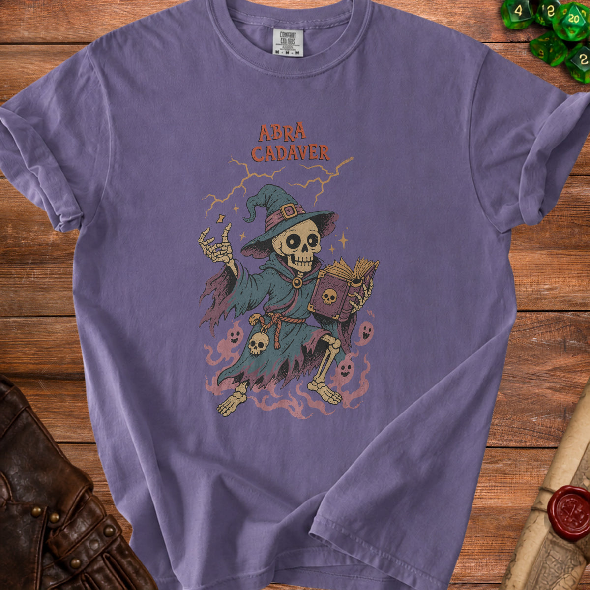 Purple t-shirt featuring skeleton wizard with book and text "Abra Cadaver."