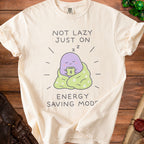 Cream t-shirt with purple character drinking coffee, text "Not Lazy Just On Energy Saving Mode".