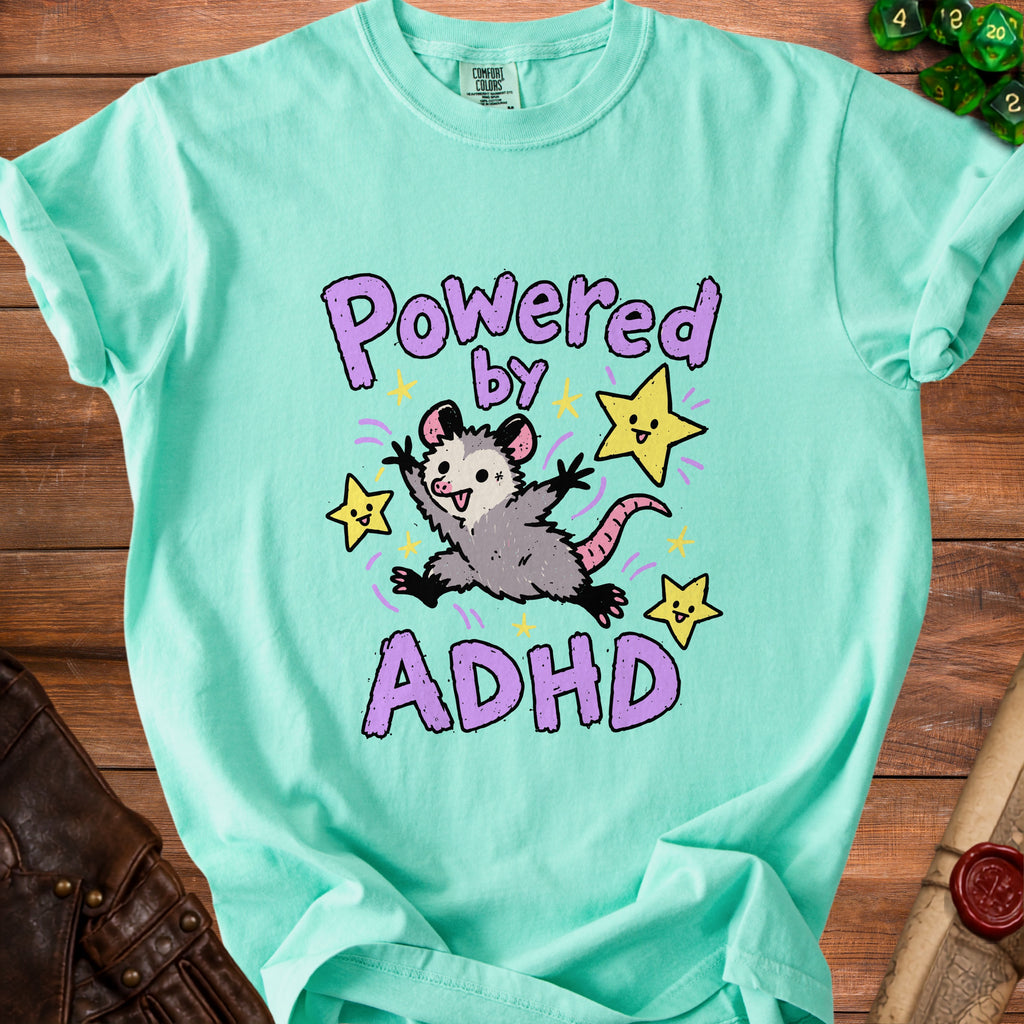 Green t-shirt with a cartoon opossum and stars graphic that says "Powered by ADHD".