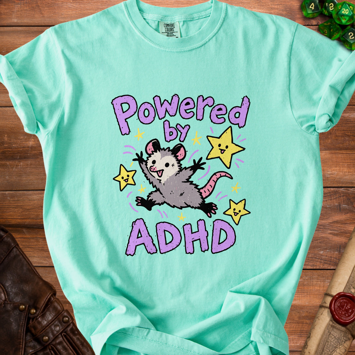 Green t-shirt with a cartoon opossum and stars graphic that says "Powered by ADHD".