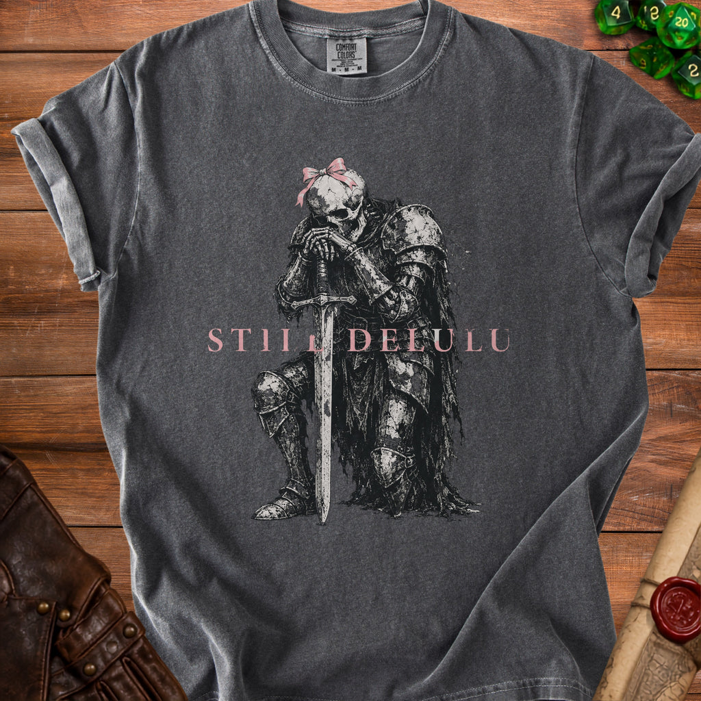 Dark grey t-shirt with skeleton knight, sword, and pink bow, "STILL DELULU" text.