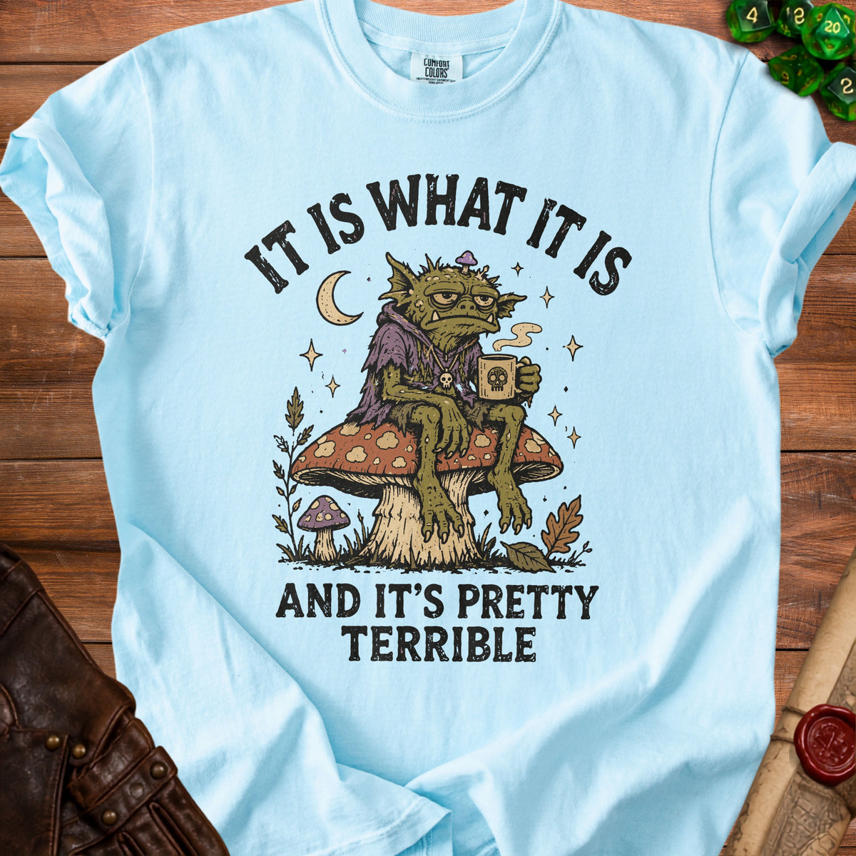 Light blue t-shirt with goblin on mushroom graphic, text "IT IS WHAT IT IS AND IT'S PRETTY TERRIBLE".