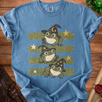 Three frogs wearing witch hats on a blue t-shirt with green stripes and stars.