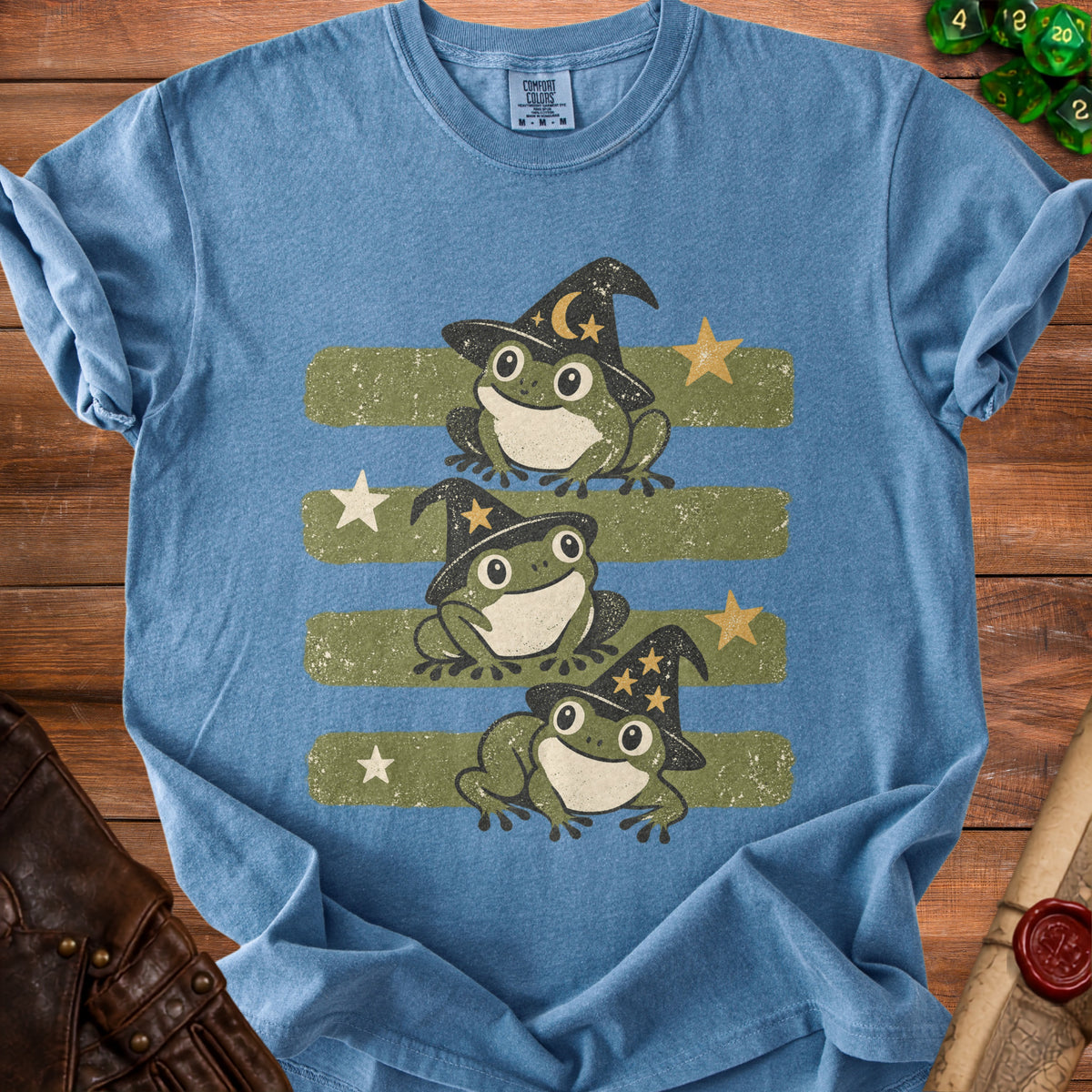 Three frogs wearing witch hats on a blue t-shirt with green stripes and stars.