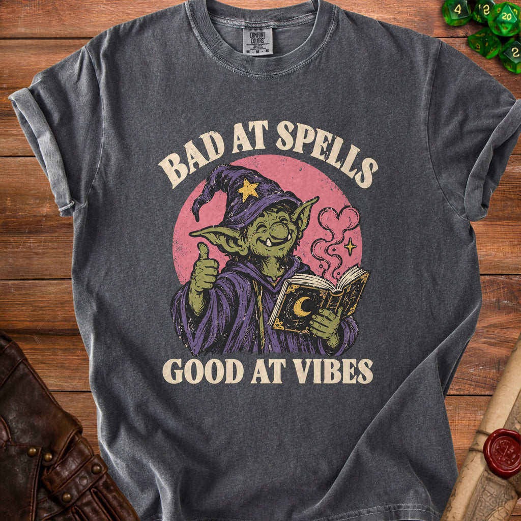 Dark gray t-shirt with graphic of goblin reading book, text "BAD AT SPELLS GOOD AT VIBES".