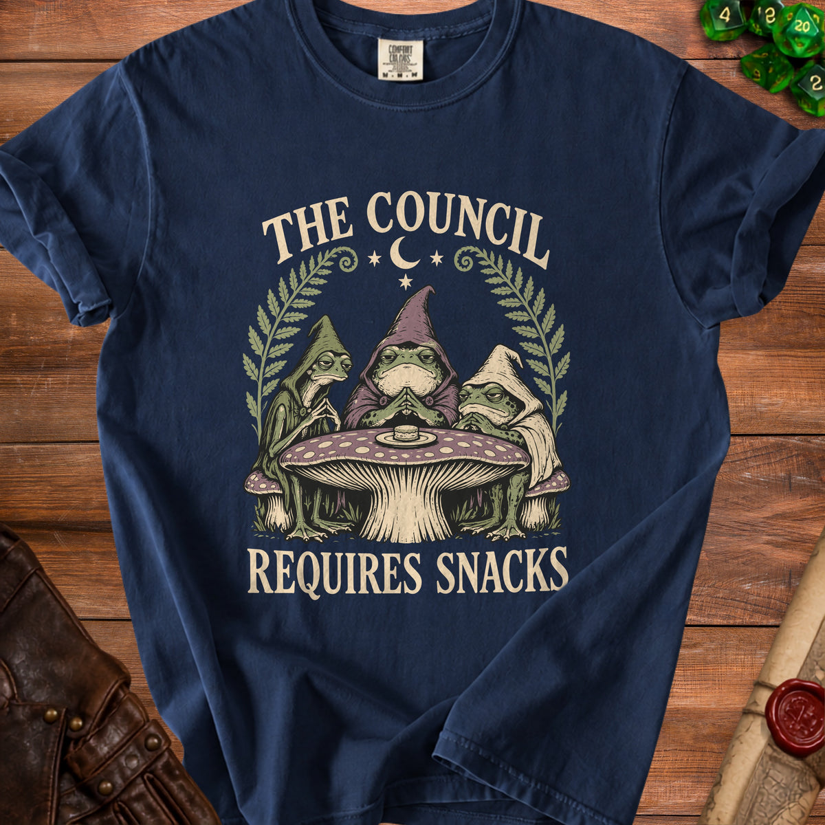 Dark blue t-shirt with graphic of three frogs wearing cloaks and hats around a mushroom table, reading "The Council Requires Snacks".