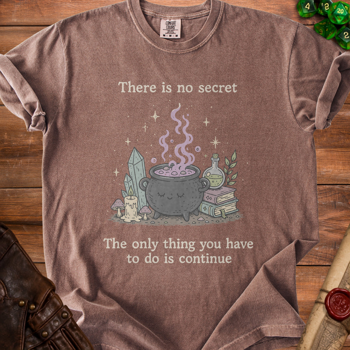 Brown t-shirt with a cauldron and crystals, reads "There is no secret The only thing you have to do is continue."
