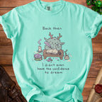 Teal t-shirt with a drawing of a creature writing and the text "Back then I didn't even have the confidence to dream".