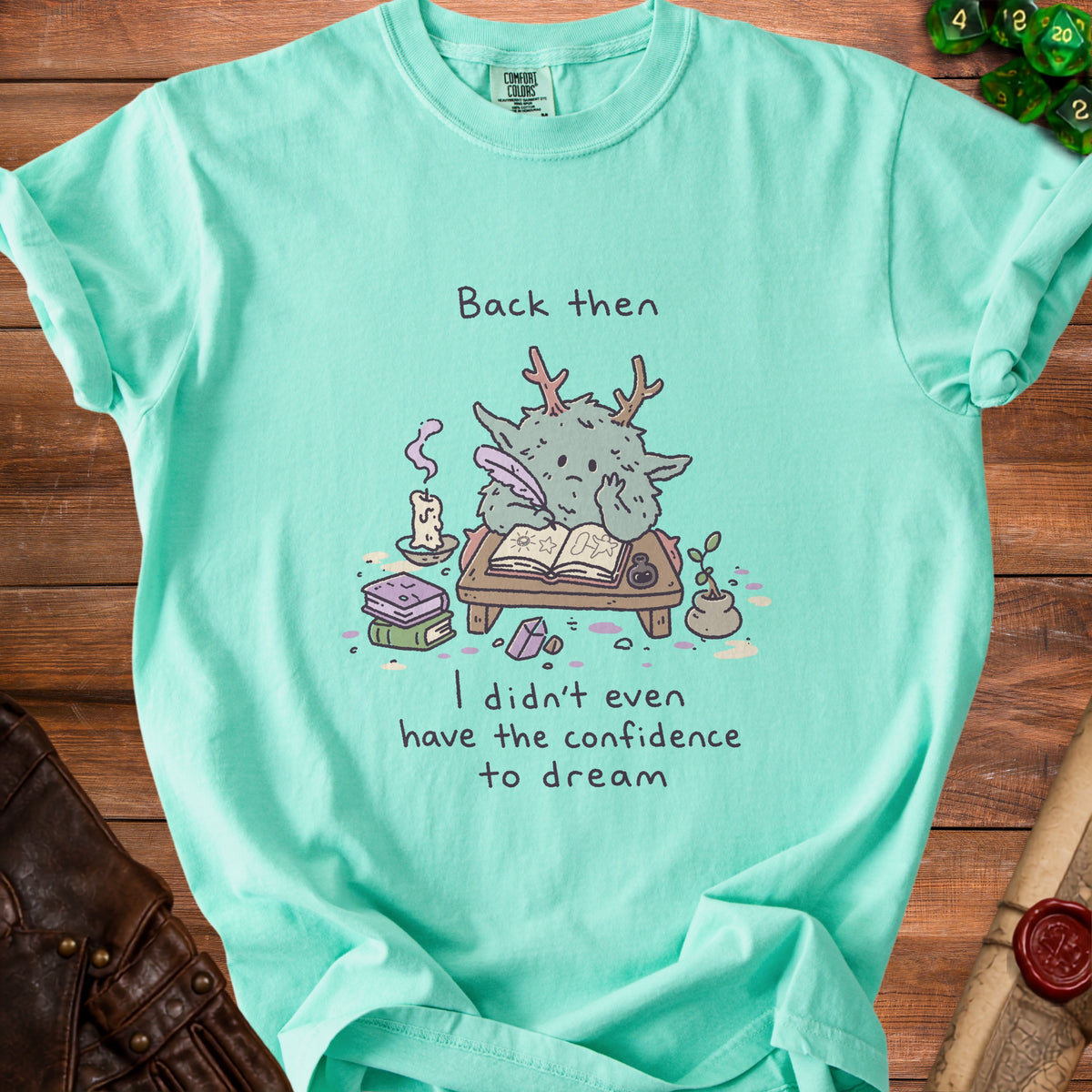 Teal t-shirt with a drawing of a creature writing and the text "Back then I didn't even have the confidence to dream".