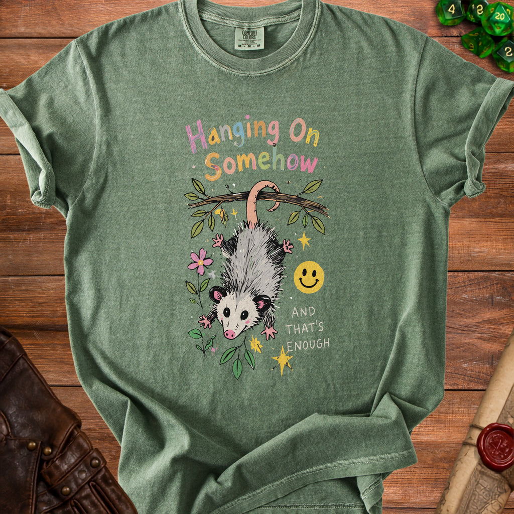 Green t-shirt with a graphic of a possum hanging from a branch, text "Hanging On Somehow" and "And That's Enough".