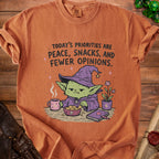 Orange t-shirt with cartoon witch eating snacks and text "Today's Priorities Are Peace, Snacks, and Fewer Opinions."