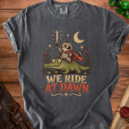 Graphic tee featuring a raccoon in a cape riding an alligator, with text "We Ride At Dawn."
