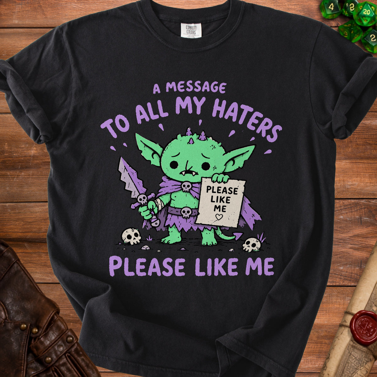 Black t-shirt with a sad green goblin holding a sign that says "Please Like Me".