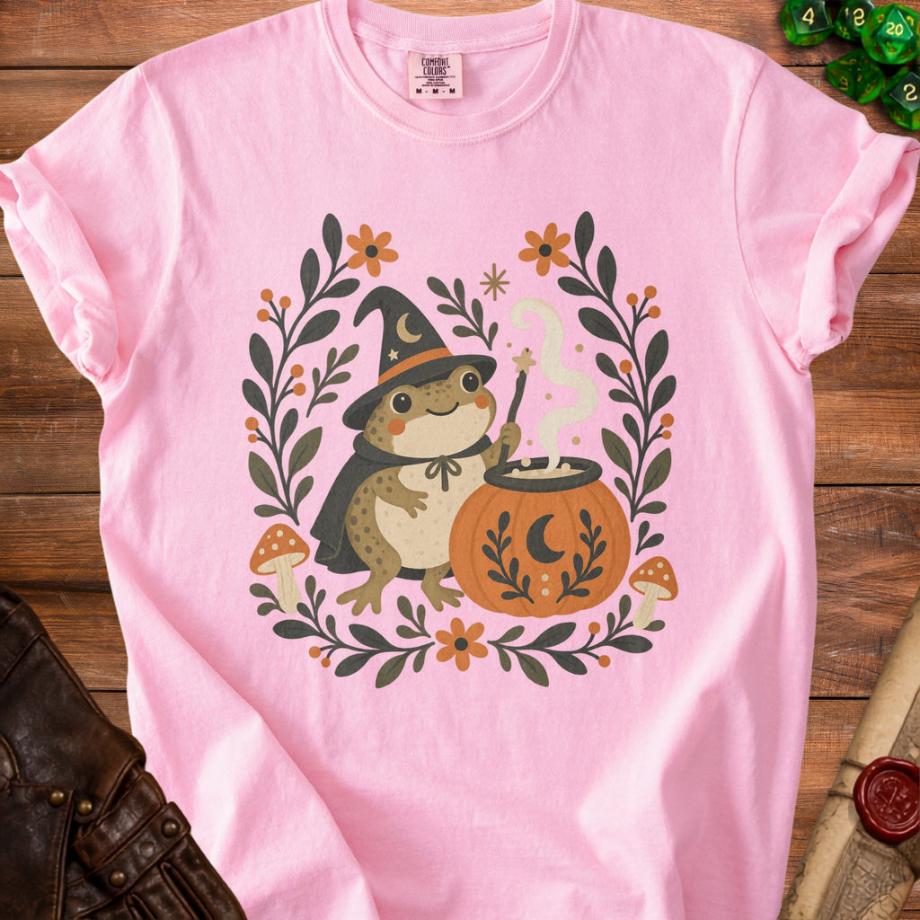Pink t-shirt with a smiling frog wizard casting a spell on a cauldron.