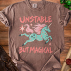 Brown t-shirt with a mint green unicorn and pink text "UNSTABLE BUT MAGICAL."