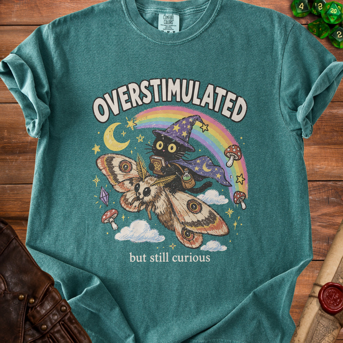Teal t-shirt with graphic of a black cat flying on a moth with text "OVERSTIMULATED but still curious".